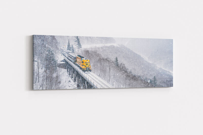 A bright yellow locomotive pulls passenger cars across a snow-covered railroad trestle bridge through a misty winter mountain landscape at Willey Brook Bridge in Crawford Notch State Park, with frost-laden trees and steep mountainsides visible through falling snow.