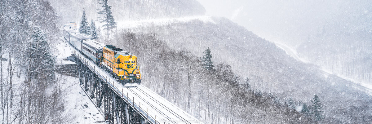 A bright yellow locomotive pulls passenger cars across a snow-covered railroad trestle bridge through a misty winter mountain landscape at Willey Brook Bridge in Crawford Notch State Park, with frost-laden trees and steep mountainsides visible through falling snow.