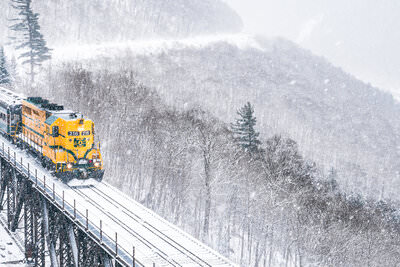 A bright yellow locomotive pulls passenger cars across a snow-covered railroad trestle bridge through a misty winter mountain landscape at Willey Brook Bridge in Crawford Notch State Park, with frost-laden trees and steep mountainsides visible through falling snow.