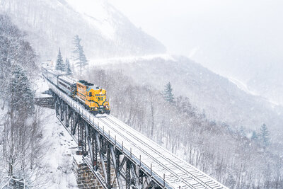 A bright yellow locomotive pulls passenger cars across a snow-covered railroad trestle bridge through a misty winter mountain landscape at Willey Brook Bridge in Crawford Notch State Park, with frost-laden trees and steep mountainsides visible through falling snow.