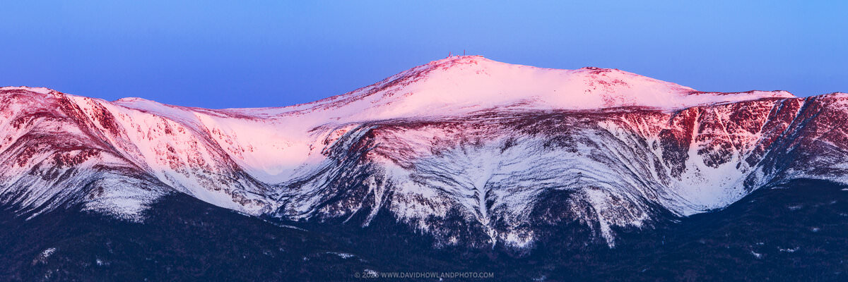 A panoramic sunrise photograph of snow-covered Mount Washington, with Tuckerman and Huntington Ravines visible as dramatic bowl-shaped glacial cirques on the mountain's face, bathed in warm pink and red alpenglow light against a deep blue pre-dawn sky, with small structures visible on the summit and dark evergreen forest in the foreground.