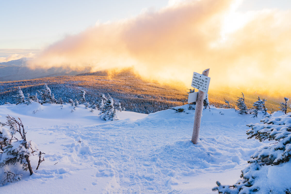 A snow-covered trail junction on the Webster Cliff Trail features a wooden signpost with directional markers, surrounded by frost-laden evergreen trees, with a dramatic golden sunset illuminating billowing clouds over a snow-dusted mountain ridge in the background.
