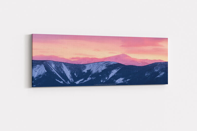 A panoramic winter landscape photograph showing snow-dusted dark forested ridgelines in the foreground, with the snow-covered Presidential Range including Mount Washington rising behind them, all bathed in vivid pink and orange alpenglow from a sunrise or sunset sky.