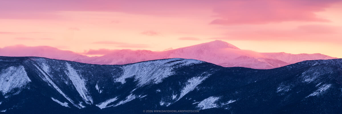 A panoramic winter landscape photograph showing snow-dusted dark forested ridgelines in the foreground, with the snow-covered Presidential Range including Mount Washington rising behind them, all bathed in vivid pink and orange alpenglow from a sunrise or sunset sky.