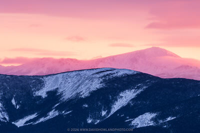 A panoramic winter landscape photograph showing snow-dusted dark forested ridgelines in the foreground, with the snow-covered Presidential Range including Mount Washington rising behind them, all bathed in vivid pink and orange alpenglow from a sunrise or sunset sky.