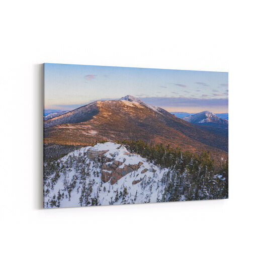 A winter sunrise vista from a snow-covered mountain summit shows rocky outcrops with scattered evergreen trees in the foreground, overlooking distant snow-dusted peaks of the White Mountains under a pastel sky with soft clouds.