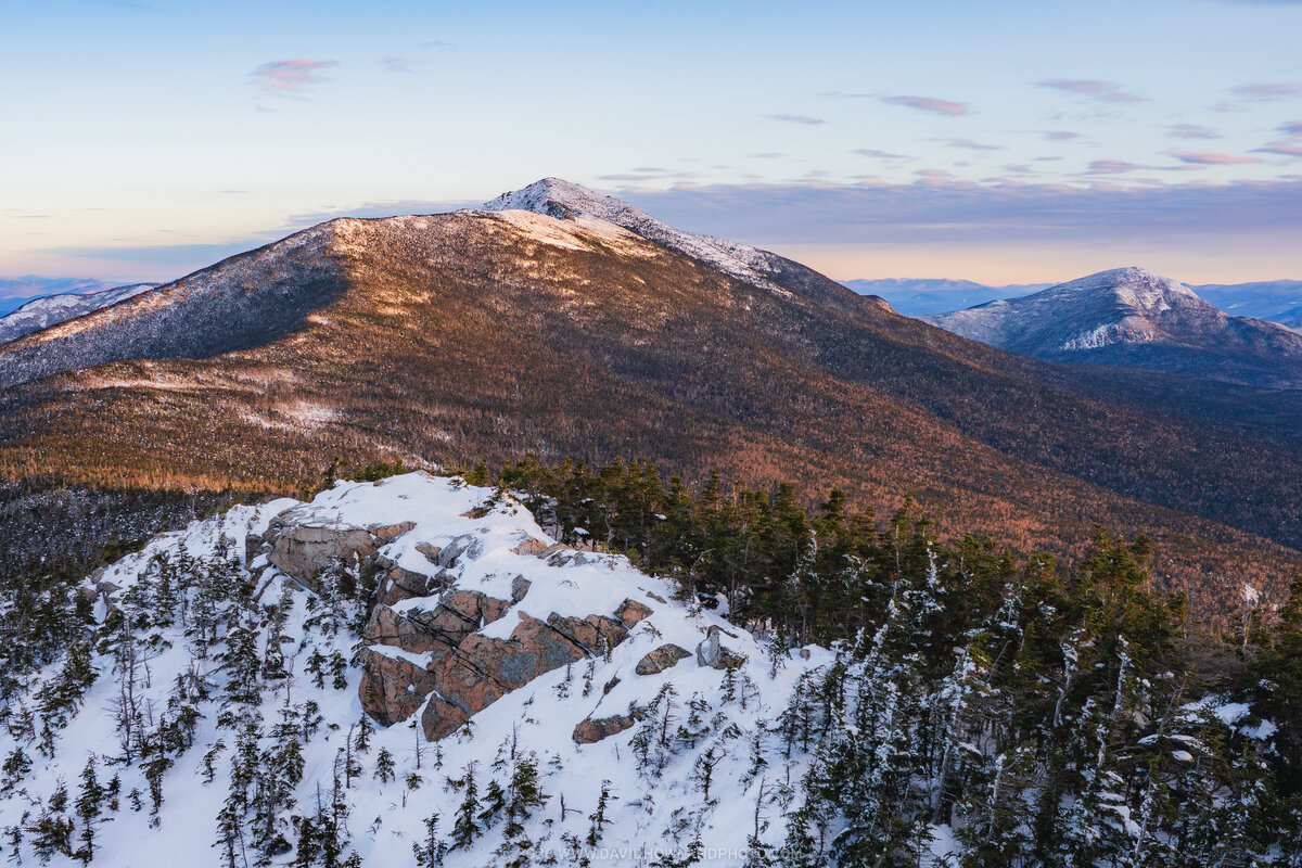 A winter sunrise vista from a snow-covered mountain summit shows rocky outcrops with scattered evergreen trees in the foreground, overlooking distant snow-dusted peaks of the White Mountains under a pastel sky with soft clouds.