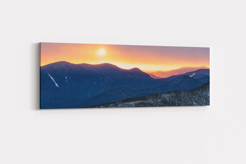 A panoramic winter sunrise photograph showing snow-dusted Mount Carrigain and surrounding White Mountain ridgelines silhouetted in deep blue, with a brilliant golden-orange sun fully risen in the upper left against a warm peach and lavender sky fading to distant purple layers of mountains on the horizon.