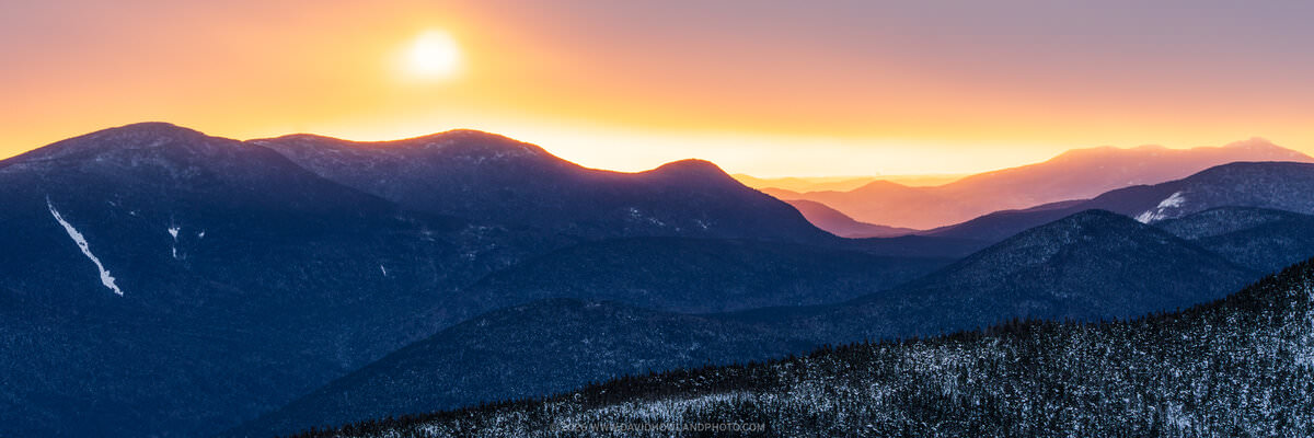 A panoramic winter sunrise photograph showing snow-dusted Mount Carrigain and surrounding White Mountain ridgelines silhouetted in deep blue, with a brilliant golden-orange sun fully risen in the upper left against a warm peach and lavender sky fading to distant purple layers of mountains on the horizon.