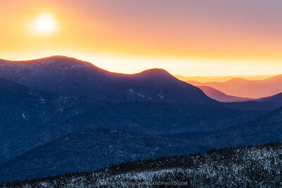 A panoramic winter sunrise photograph showing snow-dusted Mount Carrigain and surrounding White Mountain ridgelines silhouetted in deep blue, with a brilliant golden-orange sun fully risen in the upper left against a warm peach and lavender sky fading to distant purple layers of mountains on the horizon.