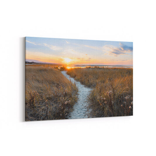 A sandy path winds through golden-brown marsh grasses toward a winter sunset over Rock Harbor Beach in Orleans, Massachusetts, with the low sun casting warm light across shallow tidal flats and distant figures visible on the shoreline.