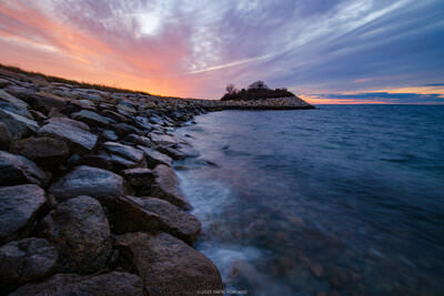 A photo of a colorful sunset behind the rocky outcropping of the Knob in Woods Hole, Massachusetts on Cape Cod.