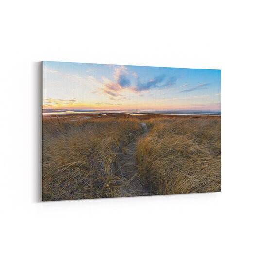 A winter sunset at Rock Harbor Beach in Orleans, Massachusetts, showing golden marsh grasses in the foreground, tidal flats stretching to the horizon, and a dramatic sky with pink, orange, and blue clouds illuminated by the setting sun.