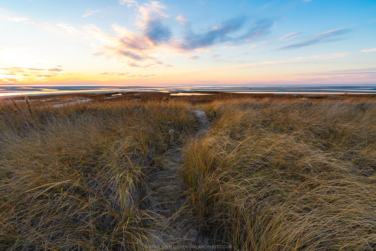 A winter sunset at Rock Harbor Beach in Orleans, Massachusetts, showing golden marsh grasses in the foreground, tidal flats stretching to the horizon, and a dramatic sky with pink, orange, and blue clouds illuminated by the setting sun.