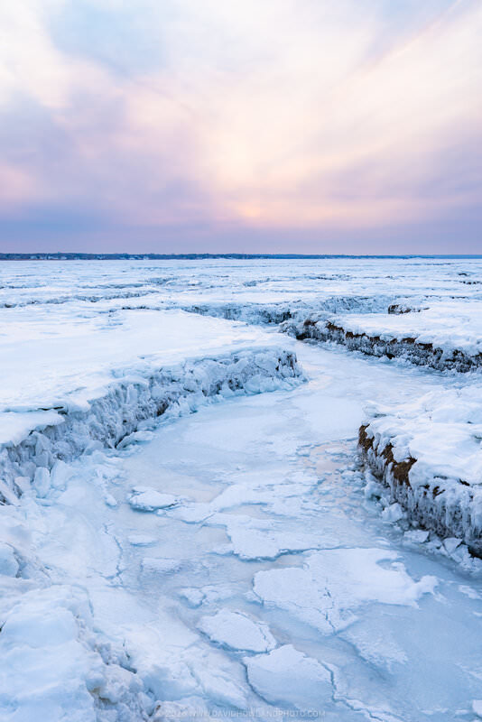 A frozen salt marsh at Bass Hole covered in ice and snow creates winding channels and textured patterns across the landscape, with a soft pink and blue sunset sky reflected in the icy terrain stretching toward the distant shoreline.
