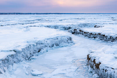 A frozen salt marsh at Bass Hole covered in ice and snow creates winding channels and textured patterns across the landscape, with a soft pink and blue sunset sky reflected in the icy terrain stretching toward the distant shoreline.